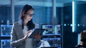 Young Female IT Employee Wearing Glasses Uses Tablet in System Control Center. In the Background Her Coworkers are at Their Workspaces with many Displays Showing Valuable Data.