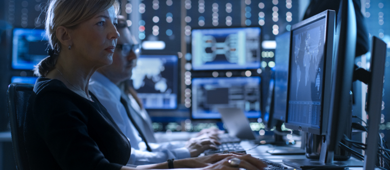 Female Government Employee Works in a Monitoring Room. In The Background Supervisor Holds Briefing. Possibly Government Agency Conducts Investigation.